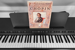Czarno-biały obraz przedstawiający pianino a na nim zbiór nut Szopena w kolorze muszlowym. Na zbiorze jego kolorowy portret