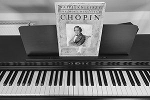 Czarno-biały obraz przedstawiający pianino a na nim zbiór nut Szopena. Na zbiorze jego portret